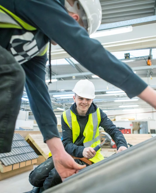 A roofing student grinning while working with a teammate to carefully install roof tiles on a sloped wooden training structure. A roofing student grinning while working with a teammate to carefully install roof tiles on a sloped wooden training structure.